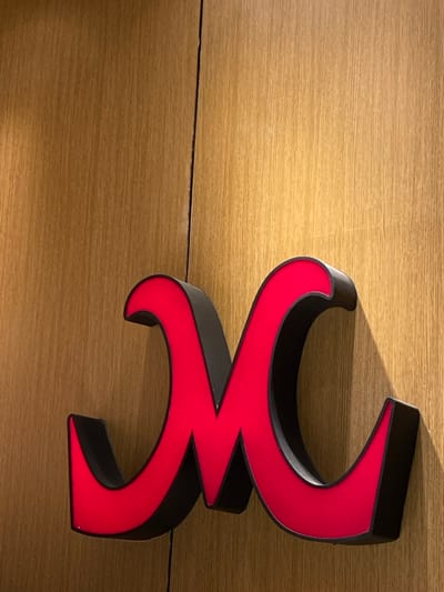 The image displays a three-dimensional, stylized letter or symbol in bright red and black, mounted on a light-colored wooden surface. The symbol features a complex, symmetrical design, resembling two mirrored '3' shapes stacked vertically, or possibly a stylized 'W' or 'M'. The front surface of the symbol is a vibrant red, while its thick, curved sides are black, giving it a prominent, dimensional appearance.

A distinct shadow is cast by the symbol onto the wooden background, extending to the upper left, suggesting an off-camera light source from the right. The background is composed of light brown wood paneling, characterized by a fine horizontal grain and a visible seam or joint running across the middle of the image. The setting appears to be indoors. There are no people or discernible activities taking place. The object functions as a decorative element or part of a logo or signage. No legible text is present. While the image is from Bekasi Regency, Indonesia, no specific visual cues within the photo directly indicate this location.