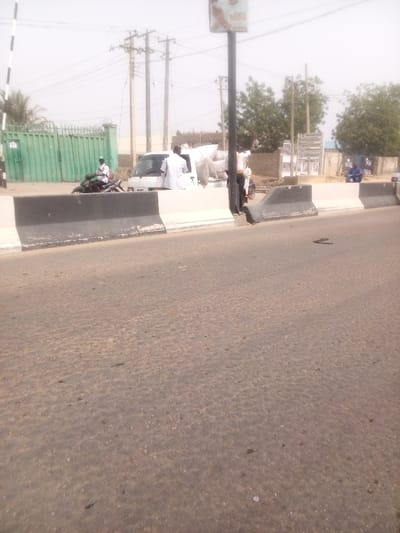 The image depicts a roadside scene in Sabon Gari East, Nigeria, under bright, possibly hazy daylight conditions. The foreground consists of a worn asphalt road surface. Two concrete barriers, painted white with black tops, run horizontally across the midground, separating the road from an adjacent area. A dark sandal or flip-flop lies on the asphalt to the right of these barriers.

Beyond the barriers, from left to right: a large green metal gate, partially open, is visible with a striped barrier arm extending from it. A dark motorcycle is positioned in front of the gate, with a male individual wearing a light garment and a cap seated on it. To the right of the motorcycle is a white utility vehicle, possibly a minivan or small truck, with a license plate "AA 487 YY." The vehicle's rear cargo area is loaded with multiple sacks. A male individual in a white garment stands by the passenger side of this white vehicle.

Further to the right, a tall black pole with an advertisement or sign mounted at its top extends upwards. Another male individual, wearing a yellow shirt and dark trousers, stands behind this pole with his right arm raised. In the background, multiple wooden utility poles with numerous overhead power lines are visible. Sparse trees and light-colored buildings are present throughout the background. A male individual in a blue garment is seated on a low wall or structure on the far right, where a sign partially reads "NGK DAVID."