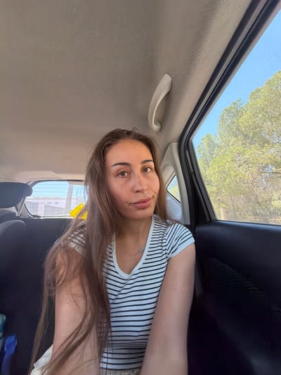 A young woman with long brown hair, wearing a black and white striped t-shirt and a silver necklace, sits in the backseat of a car. The woman is looking directly at the camera with a neutral expression. The interior of the car is visible, including the ceiling, a grab handle, and the dark seats. Through the windows, green trees and a bright blue sky are seen, suggesting an outdoor setting. The background through the rear window shows a building complex and a yellow sign. The image appears to be taken in Larnaca, Cyprus, based on the typical car interior and Mediterranean vegetation.