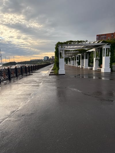 The image depicts a wet asphalt pathway adjacent to a white pergola structure covered in greenery. The pathway reflects the overcast sky and the light from the setting sun, creating streaks of light and dark. The pergola, with its repeating white columns and overhead structure, stretches into the distance along the pathway. Behind the pergola, a red brick building and trees can be seen, suggesting an urban or park setting.
In the distance, a city skyline with modern buildings is visible under a dramatic sky filled with clouds that are illuminated by the golden light of sunset. A decorative black metal fence with intricate patterns runs along the edge of the pathway, separating it from a lower level or body of water, though this is not clearly depicted.
The overall impression is one of a tranquil, possibly post-rain scene, with the wet surfaces enhancing the colors and reflections. The time of day is clearly late afternoon or early evening, indicated by the low sun angle and the golden light. The weather appears to be clearing after rain, with clouds still present but allowing sunlight to break through. The location cues, such as the architecture and skyline, combined with the provided location context of Pavlodar, Kazakhstan, place the scene in a city environment. There is no visible text in the image.