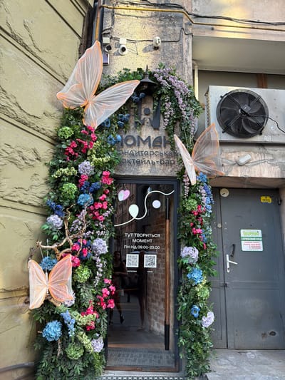 This media file captures an exterior view of a building, likely a business in Lviv, Ukraine. The scene is adorned with a lush garland of artificial greenery and vibrant flowers in shades of pink, blue, and purple, framing a window and a doorway. Delicate, translucent butterfly decorations are strategically placed, adding a whimsical touch to the facade.
Above the floral display, a large industrial fan is mounted on a grey panel. Several pieces of electrical equipment and wiring are visible on the textured stone wall to the left. The window itself reflects an interior space, hinting at a brick wall and possibly furniture.
Visible text on the door includes a green sign with Ukrainian writing, indicating "SERVICE ENTRANCE" and listing operating hours: "Goods Reception 8:00-16:00, Entrance 8:00-12:00, Weekend." Other text on the door is partially obscured. Inside the window, text in Cyrillic reads "HERE THE MOMENT IS CREATED" and provides operating hours from "Monday-Sunday 08:00-23:00." There is also a QR code and the word "MENU" and "INSTAGRAM" visible.
The overall impression is that of a charming and welcoming establishment, possibly a bar or cafe, with a playful and decorative approach to its exterior. The lighting suggests it could be daytime, with a slight shadow play indicating natural light.