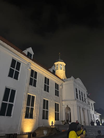 This photograph captures a colonial-era building at night, illuminated by artificial lights. The building is a white, two-story structure with dark green louvered windows and a terracotta tiled roof. A prominent octagonal tower with a clock face and weather vane rises from the center of the building. The text "GOUVERNURS KANTOOR" is visible above the entrance. Several people are scattered around the exterior of the building, some sitting on a low wall in the foreground, while others stand on the steps. One person in the foreground is taking a photo with a yellow smartphone. The sky is dark and cloudy. This building is located in Kota Tua, West Jakarta, Indonesia, and is known as the Fatahillah Museum, formerly the Governor's office.