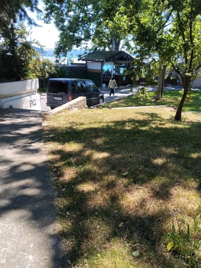 In Sarıyer, Turkey, a dark-colored van is parked on a paved area next to a stone retaining wall. A person wearing a light-colored jacket and dark pants is walking away from the van, towards a small building with a dark roof and glass windows. The building appears to be a kiosk or small shop. The ground in the foreground is covered with grass, some of which is dry and yellowed, with shadows cast by trees overhead. A concrete path runs alongside the grass. In the background, trees and distant water are visible, suggesting a waterside location. Graffiti, including the word "TAQ," is present on a white concrete structure on the left side of the image.
