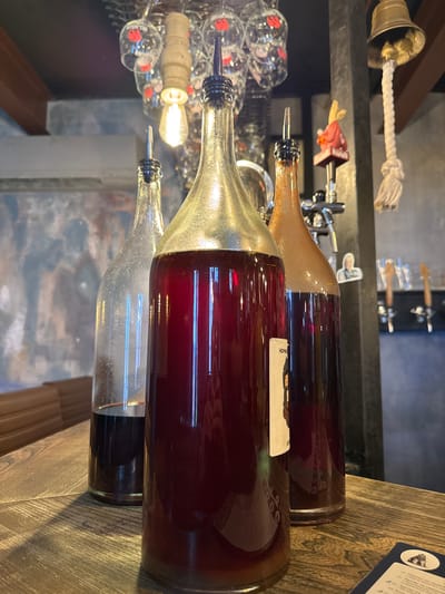 Three large glass bottles containing dark red liquid are displayed on a wooden table. The bottle in the center is the largest and has a metallic gold-colored top with a black pour spout. To its left is a clear glass bottle with a similar black pour spout, containing less liquid at the bottom. To the right of the center bottle is a slightly smaller, amber-colored glass bottle, also with a black pour spout. The background shows a bar or restaurant setting with hanging glasses, a light fixture, a bell, and a beer tap. A label with an illustration is visible on the front of the center bottle. The overall lighting is dim, with a warm tone.