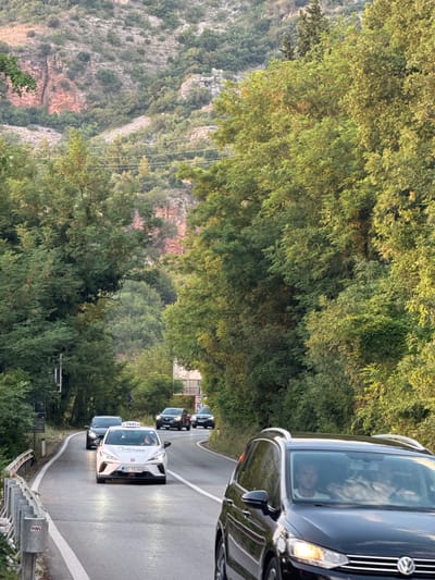 The image shows a convoy of vehicles, including cars and a van, driving along a winding road surrounded by lush green trees. The sun appears to be setting, casting a warm glow on the scene, indicating it's late afternoon or early evening. The road is paved and has white lane markings. The surrounding landscape features rocky hills and dense vegetation.

In the foreground, a white car with "TAXI" and "20" written on its windshield is prominently visible. It has a license plate that reads "BG TX066". Another car is following closely behind it. Further up the road, several other cars are visible, also driving in the same direction. A dark-colored van is at the head of the convoy.

The image was captured in Sveti Stefan, Montenegro. The overall mood is one of movement and travel, with the natural beauty of the Montenegrin landscape providing a scenic backdrop. There are no clear indications of any specific event taking place beyond a group of vehicles traveling together.