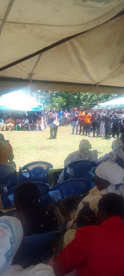 The image depicts a large group of people gathered under a large white tent. The tent is located on a grassy field. Several people are seated in blue plastic chairs, while others are standing around.  There is a man in a dark suit and tie speaking into a microphone near the middle of the group. The man is facing the people seated in the chairs. There is a large group of people standing behind the man in the suit. There are several trees in the background. The image was taken under a sunny sky.  The coordinates provided correspond to Lubao, Kenya, but it's impossible to determine the exact location within the city based on the image.