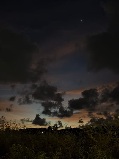 The image depicts a twilight sky with dramatic clouds. The left side of the frame is dominated by dark, ominous clouds that gradually transition to lighter hues of gray, pink, and orange towards the center, suggesting the last light of sunset or the first light of dawn. A single bright star or planet is visible in the dark sky on the left. The right side of the image features the silhouette of a dark hillside or mountain range, topped with lush, dark green foliage. The overall atmosphere is moody and somewhat dramatic, evoking a sense of mystery or anticipation. The time of day is clearly twilight, given the color of the sky and the presence of a celestial body. The weather appears to be partly cloudy, with a mix of dark and lighter cloud formations. While no people or specific objects are discernible, the scene is set outdoors with a natural landscape. The location context provided suggests the image was taken in Thep Krasatti, Thailand. There is no visible text in the image.