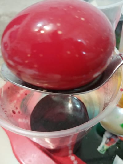 The image shows a close-up of two large, spherical objects resembling food items in a plastic cup. One is bright red and sits on a spoon above the cup, reflecting light. The other is darker and submerged inside the cup, which appears to contain a reddish liquid. There is a portion of another white, spotted object partially visible to the side of the cup. Based on the objects in the picture there is no indication of location, the city Elektrostal, Russia cannot be determined.