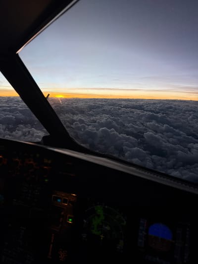 The image depicts a sunrise viewed from the cockpit of an aircraft, possibly in flight. Outside the left window, the sky transitions from a pre-dawn darkness to a bright orange horizon, partially obscured by a dense layer of clouds. A portion of the aircraft's window frame is visible. To the right of the window, the cockpit instrumentation panel is visible but largely obscured by darkness, displaying faint lights and screen readouts. The scene appears to be taken from high altitude. The location in Wanchaq, Peru, is not directly discernible from the image.