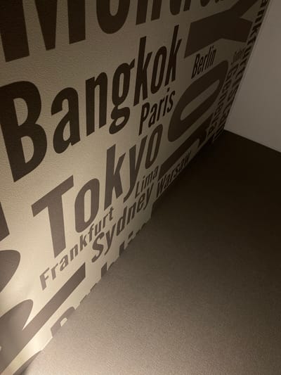 The image shows a close-up view of a wall with large, stylized text featuring various city names. The text is in a dark brown color against a light beige, textured background. Some of the visible city names include "Bangkok", "Tokyo", "Frankfurt", "Sydney", "Paris", and "Berlin". The text is arranged in a repeating pattern, with some words partially obscured or angled. The lighting suggests an indoor environment, with a shadow cast by one of the words. The overall impression is of a decorative wallpaper or mural with a travel or global theme. The location in Kamianske, Ukraine is not discernible from the visual information provided in the image.