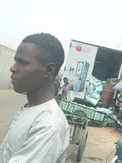 The scene depicts an outdoor street or market area in Fagge C, Nigeria, under an overcast sky. In the immediate foreground, a dark-skinned male with short, dark, curly hair is seen in left profile, wearing a light-colored, checkered long-sleeved shirt. His gaze is directed forward.
In the mid-ground, a white commercial truck, branded with the red "LG" logo and an advertisement for an LG refrigerator, is stationary with its rear cargo door open. The truck's cargo area is extensively loaded with numerous white, green, and blue plastic-wrapped packages, identified as Pampers brand diapers. Some packages are stacked within the truck, while others are piled on the ground directly behind it.
Adjacent to the truck, a man in a striped shirt is positioned, handling one of the diaper packages, appearing to either load or unload it onto a two-wheeled pushcart. The cart itself is also partially loaded with similar diaper packages. Another male figure is partially visible further back, near the rear of the truck. A brown barrel stands near the stacked packages on the ground.
The ground surface consists of dirt or unpaved earth. To the far left, a light-colored building with patterned relief is visible. On the far right, parts of a motorcycle are discernible.