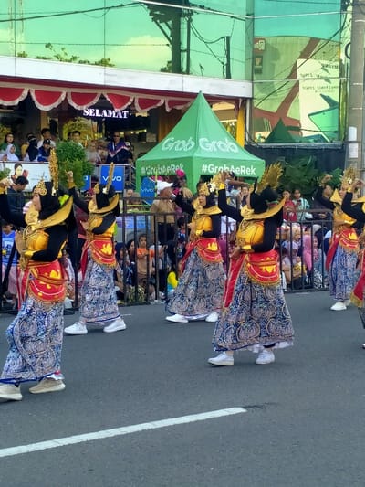 The image displays a public street scene in Madiun, Indonesia, during what appears to be a parade or cultural performance. In the foreground and midground, a line of five female individuals, identified as dancers, are in motion, positioned on an asphalt road featuring a white dividing line. Each dancer wears a costume consisting of a black long-sleeved top, a bright yellow/gold sleeveless bodice, a red and gold patterned hip sash, a long blue skirt with intricate white and gold patterns, an ornate gold headdress, and white athletic sneakers. Their arms are raised in stylized dance poses.

Behind the dancers, a crowd of spectators, varying in age, is gathered behind black metal barriers, observing the event. Some spectators are visible on a raised platform or balcony to the left. In the background, two buildings are prominent: one features a green reflective glass facade, while the other has a white facade with red and white scalloped trim and an illuminated LED sign displaying "SELAMAT." A green 'Grab' branded pop-up tent, also featuring the text '#GrabAeRek', is situated on the right side of the street. The scene is illuminated by natural daylight.