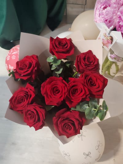 A bouquet of red roses is arranged with eucalyptus leaves in a beige paper wrap, placed on a white spherical vase decorated with black bow motifs. The bouquet is situated on a light-colored wooden surface. In the background, a large, pale peach balloon and a portion of a pink balloon with a white floral pattern are visible. To the right, a clear glass vase contains green stems and a hint of pink ribbon, accompanied by a cluster of pink peonies. A small decorative card featuring a cartoon couple is attached to a clip above the glass vase. Behind the bouquet, a dark green fabric is draped. The scene is likely an indoor setting.