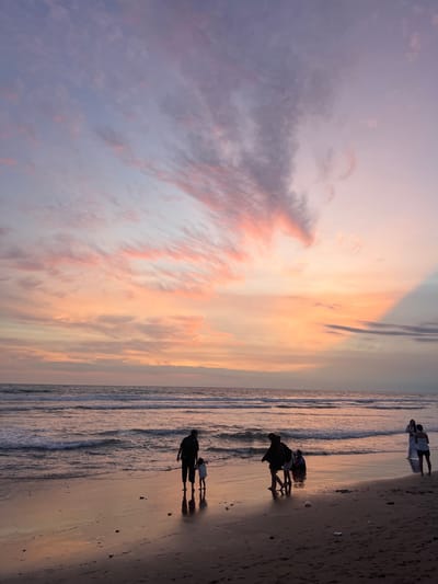 This image depicts a coastal scene at sunset on a beach in Kerobokan Kelod, Indonesia. The sky dominates the upper two-thirds of the frame, displaying stratified clouds with distinct orange, pink, purple, and pale blue hues, indicative of twilight conditions. The ocean occupies the mid-ground, featuring multiple parallel wave fronts breaking and receding on the shore. The wet, dark sand of the beach reflects the sky's colors, particularly in the foreground and mid-ground.
Six human figures are present on the beach, silhouetted against the ambient light. On the left-center, an adult figure, in dark clothing, walks leftward, holding the hand of a small child in light clothing. Slightly behind them, another adult figure, also in dark clothing, stands near the water's edge. Further back and to the right of this group, a fourth individual is partially submerged or seated in the shallow water. On the right side of the frame, two adult figures stand closer to the water. One figure, in a light-colored dress, faces towards the left. The second figure, in dark shorts and a light top, stands slightly behind and to the right of the first, also facing generally left. Scattered dark debris is visible on the sand. No overt interactions between the groups are discernible.