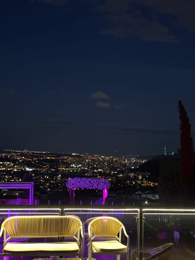 The image shows a nighttime view from a balcony overlooking the city of Mukhatskaro, Georgia. In the foreground, there are two wicker chairs with light-colored cushions. The chairs are positioned on a balcony with a glass railing that has embedded white LED lights. Behind the chairs, a vase filled with purple flowers is visible, along with other decorative elements that appear to be illuminated with purple light.
The city lights of Mukhatskaro stretch out across the landscape in the background, creating a vibrant urban glow against the dark night sky. The sky is mostly dark blue with some faint clouds. To the far right, a tall, dark, conical tree stands silhouetted against the sky. In the distance, a tower with green lights is visible on a hilltop. The overall scene suggests an outdoor seating area or restaurant with a panoramic view of the city at night.