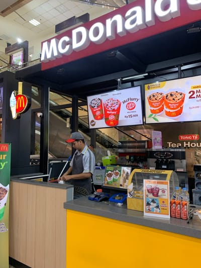 The image shows a McDonald's kiosk in a shopping mall. The kiosk is located in Malang, Indonesia at the coordinates -7.957 latitude and 112.619 longitude. The kiosk is brightly lit and features a yellow and red color scheme. There are several signs advertising McDonald's products, including McFlurries and ice cream. A man in a black shirt and a black apron is standing behind the counter, holding a cleaning tool. He appears to be cleaning the counter. There is a small sign above him that reads "McDonalds". The counter is made of gray granite and there are various food items on display. There are also multiple advertisements for different drinks and dessert items, one of which is for a tea house. A large sign above the kiosk reads "McDonald's" in large white letters. The sign is red with a black border. There are escalators in the background. The kiosk is in a well-lit, modern shopping mall.
