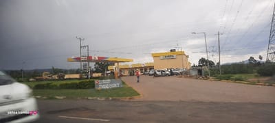 A gas station under a cloudy sky shows an Agol and Eneos business. A person is walking by the gas pumps, and several parked cars are visible. A sign in the foreground reads "All Details Auto Spares DIGCOUNT". Power lines are in the background to the right, extending upwards. A passing vehicle is blurred on the left side of the frame. The sky is overcast with dark clouds.