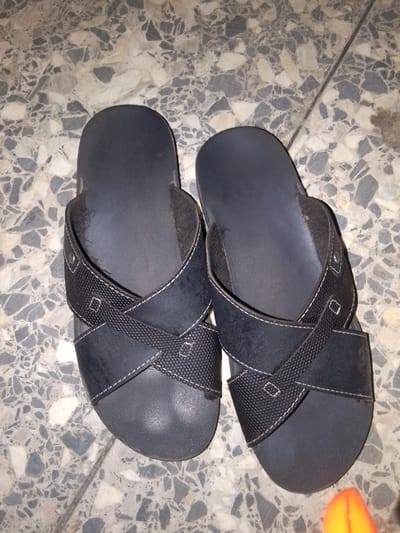 A pair of black flip-flops are displayed on a speckled gray and white tiled floor. The flip-flops feature a crossover strap design with white stitching. The left flip-flop is positioned slightly to the left and the right flip-flop to the right, both with their soles facing upwards. The floor tiles exhibit a terrazzo pattern with irregular shapes of gray, white, and black. The image is taken from a top-down perspective. This scene is likely from a residential setting within Markurdi, Nigeria, given the context.