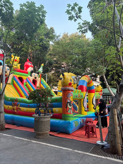 An outdoor inflatable playground, featuring characters like Pikachu, Mickey Mouse, and Angry Birds, is set up on a red matting in a park with trees. A woman in a black hijab stands to the right, watching the playground. Two red plastic stools are placed in front of the inflatable structure. The ground is paved with grey concrete. The scene appears to be in a public urban area.