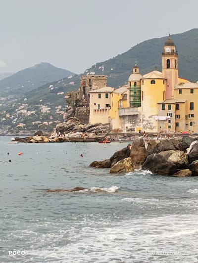 This image depicts a coastal scene in Camogli, Italy, showcasing the iconic Basilica di Santa Maria Assunta. The basilica, a prominent structure with a tall clock tower and pastel yellow and pink facade, stands on a rocky outcrop overlooking the Ligurian Sea. Multiple smaller buildings, also painted in warm, sun-bleached tones, are clustered around it, characteristic of the colorful architecture found in Italian coastal towns.

In the foreground, the sea is choppy with waves crashing against a breakwater made of large, dark rocks. Several people are visible in the water, some swimming and others wading near the shore. Others are gathered on the stone embankment and stairs leading up to the buildings. A small red inflatable raft is present in the water, and further out, buoys are scattered across the sea. The background is dominated by a lush, green hillside dotted with more houses, suggesting a densely populated coastal community extending up the slopes. The sky is overcast, casting a soft, diffused light over the scene. A digital timestamp in the lower right corner indicates the image was taken on "31.07.2026 18:29".