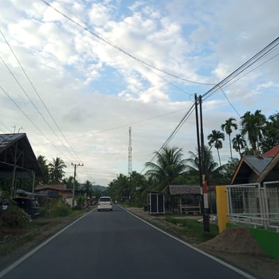 The image presents a daytime, eye-level perspective looking down a two-lane asphalt road in Uleejalan, Indonesia. The sky is partially covered with scattered cumulus clouds against a pale blue background. Numerous black overhead power lines or communication cables span across the upper portion of the sky, supported by multiple dark utility poles situated along the roadside.

On the left side of the road, in the foreground, a dark-roofed structure, possibly a shed or garage with an open side, is partially visible. A black SUV is parked beneath this structure. Further down the left, a series of buildings and houses are visible amidst dense tropical foliage, primarily palm trees. A tall, lattice-style telecommunications tower stands prominently in the mid-ground, rising above the treeline on the left side of the road.

On the right side of the road, a white multi-purpose vehicle (MPV) is traveling away from the viewer. Closer to the foreground on the right, a house with a dark, multi-gabled roof, a white metal fence, and a green wall section is visible. A pile of light-brown sand or dirt is positioned near the road edge next to this house. Further down the right, a dark, rectangular kiosk or enclosure is present, followed by several small, open-sided shelters with thatched roofs, typical of roadside stalls. Abundant palm trees and other green vegetation line both sides of the road and extend into the background, where distant hills are faintly visible. A few motorcycles are partially discernible further down the road on the right.