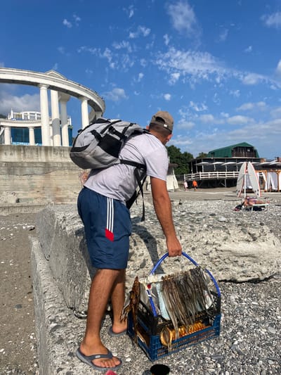 A man stands on a pebble beach in Gagra, Abkhazia, holding a blue plastic crate filled with dried fish. He is wearing a grey t-shirt, blue athletic shorts with white stripes, a beige baseball cap, and grey flip-flops. A large grey and black backpack is on his shoulders. Behind him, there is a white colonnade structure and a green building with umbrellas on a beach in the background. The sky is blue with scattered white clouds.
