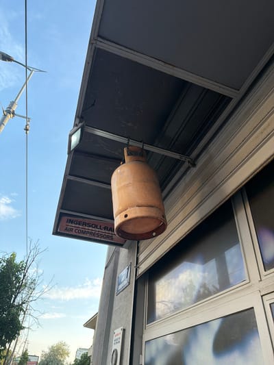 The image shows a view from below, looking up at an awning and a gas cylinder hanging from it. The awning is supported by a metal frame and appears to be attached to a building. The gas cylinder is orange and has some rust on its surface. Below the awning, there is a sign that reads "INGERSOLL-RAND AIR COMPRESSORS". The sky is blue with some clouds, and there is a street light on the left side of the image. In the distance, there are trees and buildings. The scene appears to be a street in the Palilula Urban Municipality, Serbia.