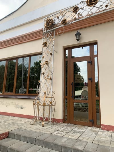 This image shows the entrance to a building in the city of Kapyĺ, Belarus. The entrance features a brown wooden door with glass panels and black hardware. Above the door, there is a decorative metalwork canopy with rose motifs. To the left of the door, there is a large window with multiple glass panes, also framed in brown wood. A decorative metal stand with floral embellishments is positioned between the door and the window. The building's exterior wall is painted a light cream color, with a decorative trim in brown and red. Steps made of grey paving stones lead up to the entrance. The ground in front of the building is paved with grey and beige interlocking stones. The reflections in the door and window show trees and a cloudy sky, and a white gazebo-like structure can be seen through the door's glass.