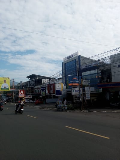 The image shows a street scene in an urban area, possibly in Zeyathiri District, Myanmar. Several buildings are visible, including a BCA bank building and a building with "Kimia Farma" signage, indicating a pharmacy. Numerous smaller signs and billboards are attached to buildings along the street. Overhead, there is a dense network of electrical wires. The road is paved and has a yellow centerline marking. A few motorcycles are driving on the road. A man is pushing a cart. The sky is partly cloudy with a mix of blue sky and white clouds. The overall setting appears to be a commercial district.