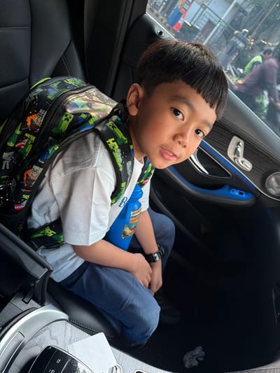 In Central Jakarta, Indonesia, a young boy with short, dark hair and bangs is seated in the back of a car. He is wearing a white t-shirt and dark blue pants, with a black backpack featuring a Minecraft-themed design adorned with green pixelated characters and orange TNT blocks. A blue water bottle with a label depicting a bird and the name "Sarah" is positioned next to him. The car's interior is visible, with black leather seats and accents of blue ambient lighting around the door panel. The boy is looking directly at the camera, his expression neutral. Outside the car, reflections in the window show a blurred street scene with vehicles and pedestrians, suggesting an urban environment.