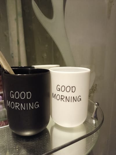 The image shows two cups, one black and one white, on a glass shelf. Both cups have "GOOD MORNING" written on them in white text. The black cup is on the left and the white cup is on the right. A toothbrush is partially visible in the black cup. The cups are in a bathroom, with a mirror reflecting a blurry image of the room. The shelf is made of glass and has a metal railing. The background is out of focus, but appears to be a tiled wall. The image does not provide any information about the location in the city of Buy, Russia.