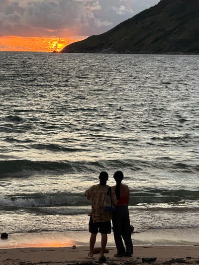 The media file shows two people standing on a beach, facing the ocean. One person, wearing a red top and dark pants, stands slightly behind and to the right of the other person. The second person, wearing a patterned shirt and dark shorts, is facing away from the camera, looking towards the water. Both individuals appear to be in silhouette against the bright water and sand.
The scene is a beach at sunset, with the sun casting a warm, golden glow on the water and sand. The ocean is calm, with gentle waves lapping at the shore. The sky is partly cloudy, with some dramatic orange and pink hues from the setting sun visible on the left side of the image, juxtaposed against a dark, steep cliff face. The overall mood is peaceful and serene, suggesting a moment of quiet contemplation or enjoyment of nature's beauty.
There is no visible text in the media file. The location cues, such as the presence of a steep cliff face meeting the sea and the warm sunset light, are consistent with a tropical coastal setting. Given the context provided, this scene is likely in Rawai, Thailand, a popular beach destination known for its scenic sunsets.