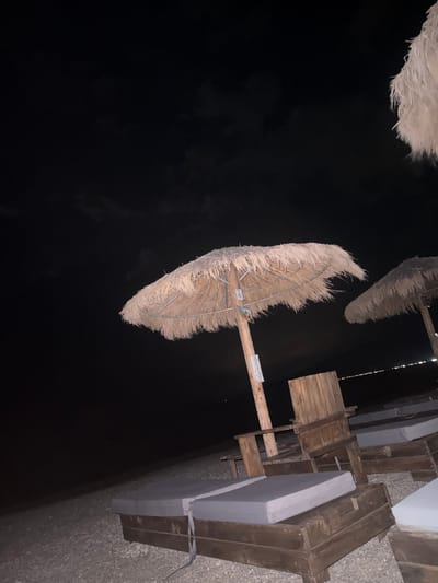 The image depicts a nighttime scene on a beach in Asprovalta, Greece. Two thatched umbrellas are visible, casting shadows on the ground. In the foreground, several wooden lounge chairs with gray cushions are arranged on the pebble-covered beach. The sea is visible in the background, with distant lights suggesting a developed area. The sky is dark, with some clouds present. The overall atmosphere is peaceful and serene, suggesting a quiet evening at the beach.