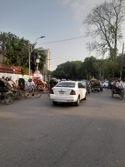 The image shows a street intersection in Dhaka, Bangladesh, at latitude 23.732 and longitude 90.395. There are several people walking and riding bicycles. A white sedan is stopped in the intersection. There is a large tree in the background. The street is paved with asphalt. There is a building on the left side of the street. The sky is overcast. There are several power lines and streetlights visible. A rickshaw with two passengers is on the left side of the road. The street is relatively busy.