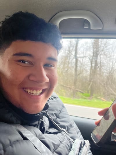 The scene is set within the interior of a vehicle, in the city of Zagore, Bulgaria. A young male with short, dark, curly hair and a light complexion is depicted from the chest up, positioned in what appears to be the front passenger seat. He is wearing a black puffer jacket, and a light-colored seatbelt crosses his chest diagonally from left to right. The individual is smiling broadly, revealing his teeth, and is looking directly towards the camera. Above the window, a light-colored grab handle is visible on the vehicle's ceiling. Through the adjacent window, a blurred outdoor environment is visible, featuring a road with a green verge on the left and numerous bare trees, indicative of a dormant season, further in the background. In the lower right corner of the frame, a hand is visible holding a black smartphone with a reflective screen. A light grey fabric pouch is partially visible near the bottom center, resting on or near the individual's lap.