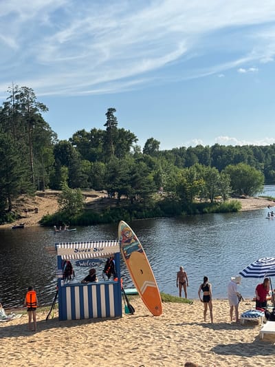 This media file depicts a sunny day at a beach by a lake, likely in Kurovskoye, Russia. The scene is bustling with activity, centered around water sports and leisure.

In the foreground, two paddleboards are visible on the sandy beach. One is orange and white, with a colorful graphic on its tail. The other paddleboard is partly obscured. Adjacent to the paddleboards is a small, blue and white striped structure, possibly a rental stand or information booth, with the text "SUPER SUP" and "Welcome" printed on it. Life jackets are also visible on and around this structure.

Several people are scattered across the beach and in the water. On the sand, a person in an orange life jacket stands near the water's edge. Further up the beach, a group of people are gathered, some sitting and others standing. One individual is wearing a white hat, and another is in a red top. Near the water, two people are lying on the sand.

In the lake, a few individuals are engaged in water activities. One person is on a paddleboard in the distance. Another group appears to be getting into or out of the water near a wooden structure.

The background is dominated by a dense forest of green trees lining the opposite bank of the lake. The sky above is a clear blue with wispy white clouds, suggesting a pleasant, warm day. The overall atmosphere appears relaxed and recreational, indicative of a summer day spent enjoying the outdoors.