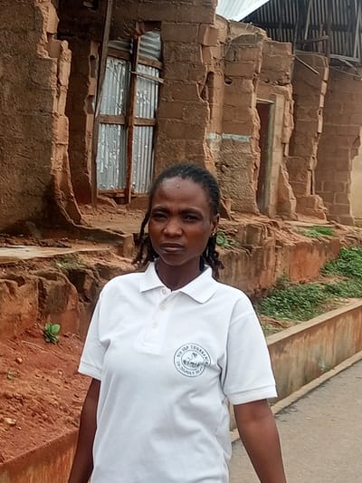 A woman with dark skin and braided hair stands in front of a dilapidated brick building. She is wearing a white polo shirt with a circular logo on the left chest. The building behind her appears to be made of reddish-brown bricks, with some sections crumbled and missing. A broken window with corrugated metal sheeting is visible. The ground in front of the building is reddish-brown soil with some green vegetation growing along a low wall. The scene is outdoors, likely in a rural or semi-urban setting in Tumfure, Nigeria.