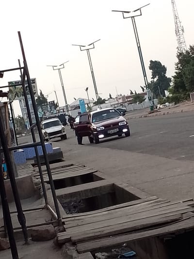 The image depicts a street scene in Pantame, Nigeria. In the foreground, a weathered wooden structure with several open rectangular pits runs along the side of the road. These pits appear to be part of a drainage system, with some showing signs of debris. A narrow paved road occupies the middle ground, with two cars driving on it. The car on the left is a white sedan, while the car on the right is a darker colored hatchback. In the background, streetlights with solar panels are visible, along with trees and buildings. A cellular tower stands prominently to the right. The sky is overcast.