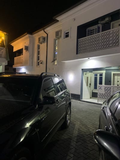 The image depicts an exterior night scene in Iranla, Nigeria, showcasing a multi-story building and two parked vehicles on an interlocking brick pavement. The building features white and dark grey/blue facades, multiple windows, and balconies with decorative white geometric pattern railings. Several wall-mounted air conditioning units are visible on the building's exterior. Artificial lights illuminate the building's facade and the paved ground. On the left, a dark-colored SUV, identifiable by its roof rails, is partially visible. On the right, the front portion of another dark-colored vehicle, likely a sedan, is partially visible. No individuals are present in the scene.