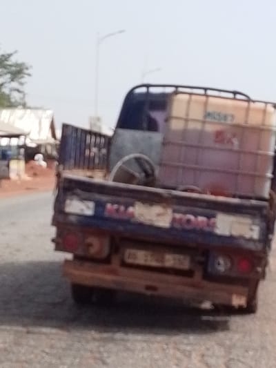 The image captures the rear view of a weathered dark-colored pickup truck, likely a Kia model, on what appears to be a road in Dungu, Ghana, during daylight hours. The truck's bed is loaded with a large, translucent off-white industrial bulk container (IBC tote) filled with a reddish-brown liquid, possibly fuel or oil. On the side of the tote, some indistinct text resembling "MESSE" is partially visible, along with a red rectangular logo or label. Also in the truck bed, a steering wheel or a similar circular object is present.

The tailgate of the truck clearly displays the faded and slightly obscured text "KIA MOTORS" in capital letters. Below the tailgate, a license plate is visible, though its alphanumeric details are too blurry to decipher. The truck's body shows signs of wear, rust, and dirt, suggesting frequent use and exposure to dusty conditions.

The setting appears to be a semi-urban or rural environment. In the background, various structures with corrugated metal roofs can be seen, hinting at shops or residential buildings. Reddish-brown earth or dirt is visible on the ground next to the road, consistent with the local terrain. A street light pole stands in the distance against a bright, possibly hazy sky. The overall impression is of a utilitarian vehicle engaged in transport in a bustling local context.