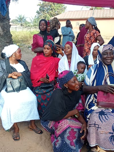 This image captures an outdoor gathering of people, predominantly women, in Doma, Nigeria, under bright daylight conditions. A red canopy or tarp is visible overhead, suggesting a planned event or temporary shelter.
In the foreground, several women are seated or squatting on the dirt ground. On the far left, a woman in a white dress, black outer garment, and white headscarf sits, holding a black handbag, with patterned sandals on her feet. Beside her, a woman in a red headscarf and a dark patterned skirt holds a mobile phone. Next, a woman in a white headscarf cradles a baby dressed in a light green and white patterned outfit. To the right, an older woman in a blue and white striped garment and patterned skirt smiles, holding a pink handbag. Another woman in a black headscarf and a vibrant patterned skirt is seen squatting low in the immediate foreground, looking to the right.
In the midground, a young woman in a black headscarf and a dark red patterned dress stands with her arms crossed, looking downwards. Other individuals, including a few men, are scattered behind the seated group. One man in a light blue tunic stands with his arms crossed. Most women wear head coverings and brightly colored, patterned traditional attire, while the men are also in traditional wear. The expressions on the faces of the subjects vary from attentive and conversational to smiling and pensive.
The background shows a simple setting with dirt ground, sparse vegetation including a prominent palm tree trunk on the left, and basic buildings with corrugated iron roofs under what appears to be a clear sky. No visible text is discernible in the image. The scene conveys a sense of community or family social interaction.
