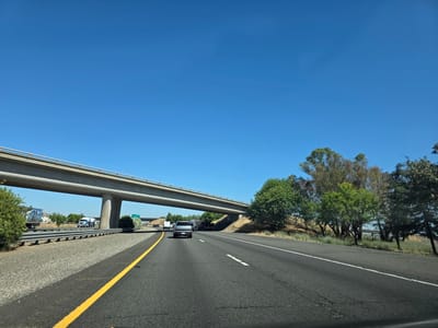This media file captures a scenic view of a highway on a clear, sunny day, likely in Sacramento, California. The perspective is from inside a moving vehicle, looking forward along the road.
The primary subjects are the highway itself, an overpass, vehicles, and a natural landscape. The highway is paved with asphalt and marked with a solid yellow line on the left and white dashed lines separating lanes. An overpass, constructed of concrete, dominates the upper left portion of the frame, supported by large pillars. Several vehicles are visible on the highway, including trucks and cars. On the left side of the highway, there's a guardrail and a gravel shoulder, with a few trees and buildings in the distance, possibly indicating a residential or industrial area. The right side of the highway features a grassy embankment with various trees, some with green foliage and others appearing drier, suggesting a warm climate.
The activity taking place is travel on the highway. The time of day appears to be daytime, given the bright sunlight and clear blue sky, with no clouds visible. The weather is fair and sunny. There are no discernible emotions to interpret, as the focus is on the setting and the act of driving. No specific text is clearly visible on road signs or vehicles to pinpoint a precise location beyond the general context of Sacramento, USA.