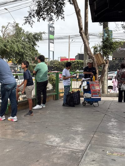 This image depicts an outdoor urban scene on a cloudy day in Santiago de Surco, Peru. The foreground shows a concrete sidewalk with cracks and discolorations. A prominent green metal fence runs from the left foreground towards the middle ground, separating the pedestrian area from what appears to be a street or commercial property.
On the left, three males are positioned near the fence. A person in a blue t-shirt and jeans is partially visible on the far left, facing away from the camera. Next to him, a young male in a dark blue t-shirt and tan shorts leans against the fence, holding a white rectangular object in his right hand. To his right, an adult male in a light green polo shirt and black pants also leans against the fence, looking towards the right side of the frame.
In the middle ground, a street vendor, an adult male wearing a light-colored hat, dark t-shirt, and dark pants, stands behind a blue, two-wheeled vending cart. The cart features a red and white display board with text in Spanish, including "PAPA RELLENA," "HUEVO," "CHAUFA," and "BEBIDAS." Several items, including plastic bottles (one black, one yellow/orange) and white bags, are arranged on the cart. The vendor is interacting with a male customer in a white t-shirt and jeans, who is holding a white food container. A black and plaid backpack rests on the ground near the customer's feet.
To the right of the vendor, an adult female is mostly visible from the back, wearing a colorful patterned top and dark pants, and holding a white bag. Further into the background, a street with vehicles, including a white van, is visible.
The environment includes numerous trees and green foliage, some with exposed trunks. A tall, light brown tree trunk is centrally located behind the vendor, with a wooden crate attached to its upper portion. Overhead, utility poles and a dense network of power lines are visible against the cloudy sky. In the background, commercial signage is present, including a tall black sign reading "JOCKEY TE QUIERE FELIZ" and "JWORK," and another sign with "EL REFUGIO" partially visible.