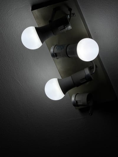 The image shows three light bulbs mounted on a fixture attached to a wall. Two of the bulbs are fully illuminated and visible, while the third is mostly obscured by shadow. The light bulbs are round and white, emitting a bright light. The fixture itself appears to be made of a metallic material and is mounted on a textured, light-colored wall. The lighting in the image is dim, with much of the scene cast in shadow, suggesting that the light bulbs are the primary source of illumination. There is some visible text on the base of the light bulbs, including "60W MAX". The setting appears to be indoors, possibly a hallway or a room with limited natural light. No people are visible in the image. The overall mood is functional and perhaps a bit stark due to the limited illumination and focus on the utility of the light fixtures. There are no overt cues to indicate the time of day, weather, or specific emotional tone beyond the functional aspect of the lighting. Given the location context of Ulan-Ude, Russia, the setting could be a residential or commercial interior.