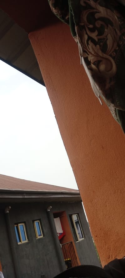The scene, located in Eziobodo, Nigeria, captures architectural elements under an overcast sky. A prominent textured, orange-brown column occupies the right and upper portions of the frame. Part of a dark green fabric with a cream-colored floral pattern hangs from an unseen point in the upper right. To the left, a multi-story building with dark grey walls is visible, featuring a red corrugated metal roof and a dark gutter system. Two grey vertical pipes with white elbow fittings are mounted on the building's facade. The building includes three rectangular windows with light frames; two exhibit reflective blue panes, and one to the right is open. Between the right two windows, a recessed orange balcony alcove contains a hanging white towel and a red fabric with a star pattern, protected by a dark metal railing. No individuals are visible.