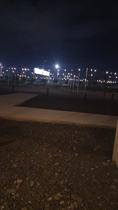 The image depicts an outdoor scene at night, characterized by a dark sky and numerous small, bright lights strung along poles, resembling festive or decorative lighting. The lights create a visual pattern stretching into the distance, suggesting an open area like a street, park, or event venue.
The foreground is dominated by a textured surface of dark gravel or small stones, with a distinct light-colored concrete barrier or curb running diagonally across the image. Behind this barrier, a dark asphalt surface is visible, likely a road or pathway.
Further back, a large, rectangular white structure, possibly a billboard or a screen, stands illuminated. This structure is also surrounded by more of the small, decorative lights. The scene suggests a public space that is lit up at night, possibly for an event or for general illumination.
There is no visible activity or people in the frame. The time of day is clearly night, with the darkness of the sky and the artificial lighting being prominent features. No specific weather conditions are apparent. The location cues, such as the concrete barrier and the gravel, along with the overhead lights and the large white sign, create a modern, urban, or semi-urban nighttime ambiance. No text is discernible in the image.