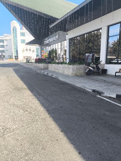 The image displays an outdoor scene in Kızılbaş, Northern Cyprus / Cyprus, featuring an asphalt road, a sidewalk, and two prominent buildings under a clear sky. A sharp diagonal shadow line bisects the asphalt road in the foreground. Along the right side, a paved sidewalk with a black and white painted curb fronts a supermarket building. The supermarket's facade includes large reflective glass windows and a prominent sign reading "the Supermarket" in white letters on a grey panel. Above the sign, a dark grey canopy, topped with a green roof section, is adorned with numerous small, dangling light elements. Below the windows, a raised concrete planter box contains green foliage. A white scooter with a dark seat and a grey and red delivery box is parked on the sidewalk near a black trash bin. A wooden bench is partially visible on the far right. In the background on the left, a multi-story white building with distinctive modern architecture, including curved elements and vertical windows, is visible. In front of this building are several large, abstract sculptures. Green trees and vegetation are present in the background. No people or active interactions are depicted.