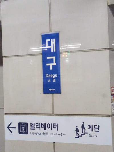 The image captures a slightly tilted view of an interior wall within a railway station in Daegu, South Korea. The walls are covered in light-colored, possibly glossy, tiles, reflecting artificial lighting.
Two prominent signs are displayed on the wall. On the left, a horizontal blue sign features the station name in multiple languages: "대구" (Korean), "Daegu" (English), and "大邱" (Chinese). A small "KORAIL" logo is also visible on this sign, identifying it as part of the South Korean national railway system.
On the right, a vertical white sign provides navigational information. The top section indicates "계단" (Korean for stairs), accompanied by the English word "Stairs" and a pictogram of two figures ascending stairs. The bottom section of this sign directs to the "엘리베이터" (Korean for elevator), with additional text "Elevator" (English), "电梯" (Chinese), and "エレベーター" (Japanese). Below this multilingual text, an elevator pictogram (a person inside a box) is shown, followed by a large downward arrow, indicating the direction of the elevator.
The scene suggests an indoor public space, likely a concourse or platform area, designed to guide passengers. No actual people are present, only the symbolic figures on the directional sign. Reflections of ceiling lights are visible on the tiled surfaces and the blue sign. The presence of multiple languages on the signs caters to both local and international travelers.