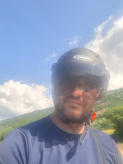 A man is wearing a black helmet with a visor, a blue shirt, and a harness with an orange clasp. The man has a beard and stubble. He is outdoors, with a blue sky and clouds in the background. There is a green, hilly landscape behind him. The text "YAWA" is visible on the side of the helmet. The image appears to be taken on a sunny day. The orientation of the image suggests the man may be paragliding or engaged in a similar activity.
