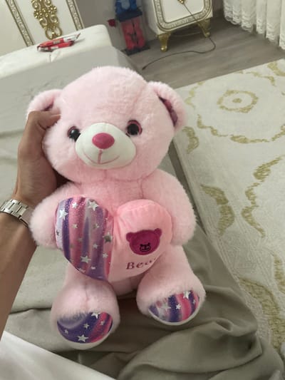 The image displays a pink teddy bear held by a person's hand in an indoor setting. The teddy bear is the central subject, made of soft, light pink plush material. It features black eyes, a white snout with a pink nose and mouth detail. Its belly and the soles of its feet are adorned with a unique fabric patterned with white stars against a gradient of pink, purple, and blue colors. On the bear's chest, a smaller pink heart with a darker outline is visible, containing the letter "B" or possibly the word "Bear" written vertically.

A human hand, appearing to be an adult's, supports the teddy bear from below. A silver-toned watch with a metallic link band is worn on the wrist.

The background suggests a cozy interior space. On the left side, there's a white surface with gold, ornate decorative accents, possibly part of a clock or furniture. Above it, a white, ruffled or lace-like curtain is partially visible. The floor or wall in the background is covered by a light-colored rug or carpet with subtle, gold-tan swirling patterns. A thin black wire or cord is also present on this surface. To the right, a greyish-tan fabric, likely a blanket or bedding, is draped. A small red toy car is faintly seen in the far left background. The overall lighting is soft and ambient, indicative of an indoor environment. No specific activity beyond holding the bear is clearly depicted. No direct visual cues indicate the location as Ihtiman, Bulgaria.